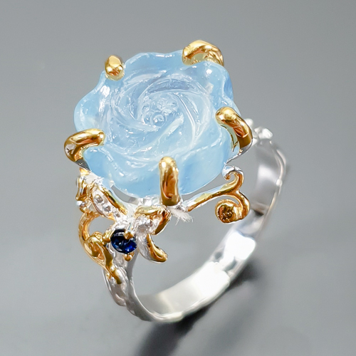 Δαχτυλίδι σε ασήμι 925 με λευκό και κίτρινο επιχρύσωμα στα 14Κ με μια σκαλιστή Ακουαμαρίνα σε σχήμα τριαντάφυλλου με Transparent καθαρότητα διαφάνεια και βάρος 11.33 ct με έντονο γαλάζιο χρώμα. Το κόσμημα συμπληρώνουν και ένα μπλε Ζαφείρι σε στρογγυλή κοπή στα 0.10 ct. Η προέλευση των λίθων είναι απο την Βραζιλία και την Μοζαμβίκη.