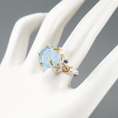 Δαχτυλίδι σε ασήμι 925 με λευκό και κίτρινο επιχρύσωμα στα 14Κ με μια σκαλιστή Ακουαμαρίνα σε σχήμα τριαντάφυλλου με Transparent καθαρότητα διαφάνεια και βάρος 11.33 ct με έντονο γαλάζιο χρώμα. Το κόσμημα συμπληρώνουν και ένα μπλε Ζαφείρι σε στρογγυλή κοπή στα 0.10 ct. Η προέλευση των λίθων είναι απο την Βραζιλία και την Μοζαμβίκη.