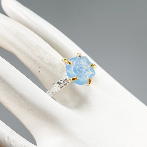 Δαχτυλίδι σε ασήμι 925 με λευκό και κίτρινο επιχρύσωμα στα 14Κ με μια σκαλιστή Ακουαμαρίνα σε σχήμα τριαντάφυλλου με Transparent καθαρότητα διαφάνεια και βάρος 11.33 ct με έντονο γαλάζιο χρώμα. Το κόσμημα συμπληρώνουν και ένα μπλε Ζαφείρι σε στρογγυλή κοπή στα 0.10 ct. Η προέλευση των λίθων είναι απο την Βραζιλία και την Μοζαμβίκη.