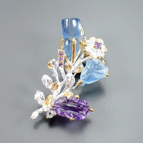 Καρφίτσα σε ασήμι 925 με λευκό και κίτρινο επιχρύσωμα στα 14Κ με ένα σκαλιστό Αμέθυστο σε σχήμα Φύλλου με transparent/Vvs καθαρότητα διαφάνεια στα 11.55 ct βάρος και έντονο μοβ χρώμα. Το κόσμημα περιέχει και δύο σκαλιστές Ακουαμαρίνες σκαλισμένη σε μορφή Κρίνων με transparent καθαρότητα στα 23.19 ct με έντονο γαλάζιο χρώμα,  τέλος το κόσμημα συμπληρώνουν και 4 αμέθυστοι σε στρογγυλή κοπή με transparent/vvs διαφάνεια καθαρότητα και συνολικό βάρος 0.35 ct. Η προέλευση των λίθων είναι απο την Βραζιλία.