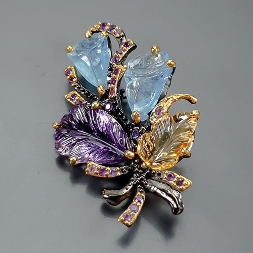 Καρφίτσα σε ασήμι 925 με μαύρο και κίτρινο επιχρύσωμα στα 14Κ με ένα σκαλιστό Αμέθυστο σε σχήμα Φύλλου με transparent/Vvs καθαρότητα διαφάνεια στα 16.27 ct βάρος και έντονο μοβ χρώμα. Το κόσμημα περιέχει και δύο σκαλιστές Ακουαμαρίνες σκαλισμένη σε μορφή Κρίνων με transparent καθαρότητα στα 29.68 ct με έντονο γαλάζιο χρώμα, Ένα Κιτρίνη Χαλαζία σκαλιστό σε σχήμα φύλλου με if-Vvs καθαρότητα και βάρος 10.09 ct και τέλος το κόσμημα συμπληρώνουν και 24 αμέθυστοι σε στρογγυλή κοπή με transparent/vvs διαφάνεια καθαρότητα και συνολικό βάρος 1.20 ct. Η προέλευση των λίθων είναι απο την Βραζιλία.