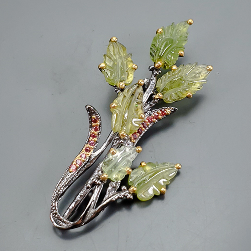 Καρφίτσα σε ασήμι 925 με μαύρο και κίτρινο επιχρύσωμα στα 14Κ με έξη σκαλιστές πράσινες Τουρμαλίνες (Βερντελίτες) σε σχήμα φύλλων με Translucent/transparent καθαρότητα διαφάνεια στα 33.60 ct βάρος. Το κόσμημα περιέχει και 14 πετράδια Ροδολίτη Γρανάτη σε στρογγυλή κοπή με vvs διαφάνεια και βάρος 0.28 ct. Η προέλευση των λίθων είναι απο την Νιγηρία και την Τανζανία.