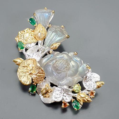 Καρφίτσα/Μενταγιόν σε ασήμι 925 με λευκό και κίτρινο επιχρύσωμα στα 14Κ με τρία πετράδια σκαλιστού Λαβραδορίτη σε σχήματα λουλουδιών με Translucent καθαρότητα διαφάνεια στα 53.88 ct βάρος. Το κόσμημα περιέχει και 4 Σμαράγδια σε κοπή Pear με transparent καθαρότητα στα 2.40 ct. Ένα Περίδοτο στρογγυλό με vvs διαφάνεια και βάρος 0.40 ct και τέλος το κόσμημα συμπληρώνουν και 2 κίτρινα Ζαφείρια σε round κοπή με vvs διαφάνεια καθαρότητα και συνολικό βάρος 0.20 ct. Η προέλευση των λίθων είναι απο την Μαδαγασκάρη, την Βραζιλία, το Πακιστάν και την Σρι-Λάνκα.