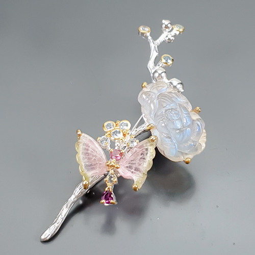 Καρφίτσα σε ασήμι 925 με λευκό και κίτρινο επιχρύσωμα στα 14Κ με ένα σκαλιστό Λαβραδορίτη σε σχήμα λουλουδιού με Translucent καθαρότητα διαφάνεια στα 25.67 ct βάρος. Το κόσμημα περιέχει και δύο σκαλιστές watermelon Τουρμαλίνες σκαλισμένες σε σχήμα φτερών πεταλούδας με transparent καθαρότητα στα 8.00 ct με έντονο Ροζ-πράσινο-λευκό χρώμα. Εννέα φυσικές Ακουαμαρίνες στρογγυλές με vvs διαφάνεια και βάρος 1.80 ct και τέλος το κόσμημα συμπληρώνουν και 2 Ροδολίτες Γρανάτες σε round+Pear  κοπή με vvs διαφάνεια καθαρότητα και συνολικό βάρος 0.40 ct. Η προέλευση των λίθων είναι απο την Μαδαγασκάρη, την Βραζιλία και την Τανζανία.