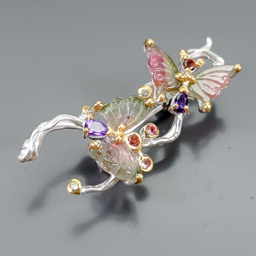 Καρφίτσα σε ασήμι 925 με λευκό και κίτρινο επιχρύσωμα στα 14Κ με σκαλιστές watermelon Τουρμαλίνες σκαλισμένες σε σχήμα φτερών πεταλούδας με transparent καθαρότητα στα 10.32 ct με έντονο Ροζ-πράσινο-λευκό χρώμα. Δύο Αμέθυστους σε  pear με vvs διαφάνεια και βάρος 1.20 ct και τέλος το κόσμημα συμπληρώνουν και 4 Ροδολίτες Γρανάτες σε round κοπή με vvs διαφάνεια καθαρότητα και συνολικό βάρος 0.40 ct. Η προέλευση των λίθων είναι απο την Νιγηρία, την Βραζιλία και την Τανζανία.