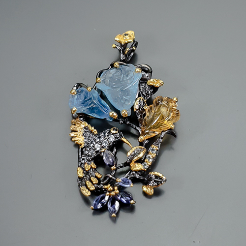 Χειροποίητο μενταγιόν σε ασήμι 925 με μαύρο και κίτρινο επιχρύσωμα στα 14Κ με δύο σκαλιστές Ακουαμαρίνες σε κοπές hand carved flower στα 25.77 ct συνολικό βάρος με transparent διαφάνεια καθαρότητα και έντονο γαλάζιο χρώμα. Στο κέντρο του κολιέ το μοτιφ αναπαριστά ένα ανοιξιάτικο τοπίο με ένα Κολιμπρί ανάμεσα σε υπέροχα λουλούδια το σώμα του κολιμπρί  ήταν κατάμεστο απο λευκά Τοπάζια  στα 0.65 ct . Δυο σκαλιστές ακουαμαρίνες σε σχέδιο Κρίνων με συνολικό βάρος 25.77 ct ένα Σκαλιστό φύλλο Κιτρίνη στα 9.50 ct  με χρυσοκίτρινο χρώμα και if  διαφάνεια καθαρότητα. Τέλος το κόσμημα περιεχέι και 5 φυσικούς Ιολίτες  σε κοπή marquise με Vvs διαφάνεια καθαρότητα και συνολικό βάρος 3.00 ct. Η προέλευση των λίθων είναι από την Βραζιλία και την Αφρική.