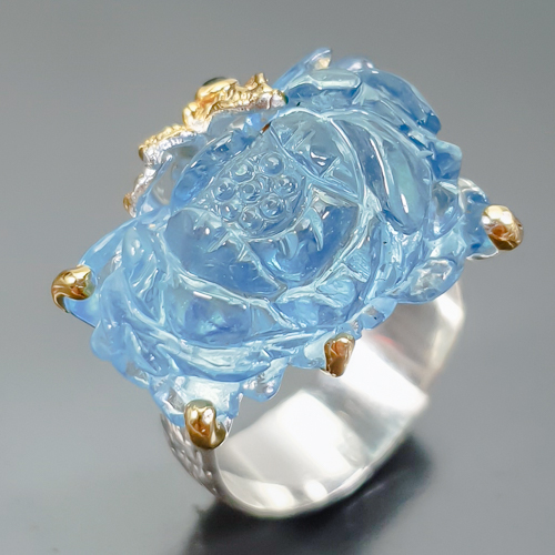 Δαχτυλίδι σε ασήμι 925 με λευκό και κίτρινο επιχρύσωμα στα 14Κ με σκαλιστή Ακουαμαρίνα σκαλισμένη σε σχήμα Λουλουδιού με if-vvs καθαρότητα στα 26.78 ct με έντονο γαλάζιο χρώμα, Το κόσμημα συμπληρώνουν και 2 ζαφείρια στα 0.04 ct σε στρογγυλή κοπή και opaque διαφάνεια. Η προέλευση των λίθων είναι απο την Βραζιλία και την Μοζαμβίκη.